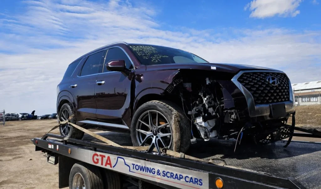 A professional flatbed tow truck from GTA Towing & Scrap Cars parked in an industrial lot with a weathered blue sedan secured on the back, featuring a backdrop of the Toronto skyline.
