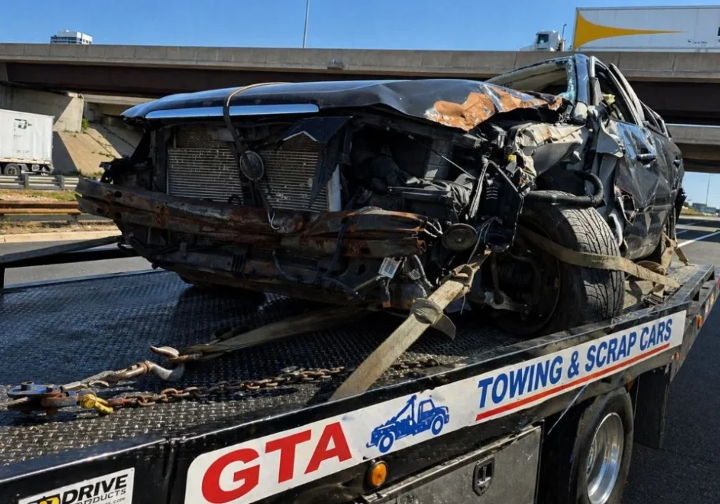 A professional flatbed tow truck from GTA Towing & Scrap Cars parked in an industrial lot with a weathered blue sedan secured on the back, featuring a backdrop of the Toronto skyline.scrap vehicle pickup