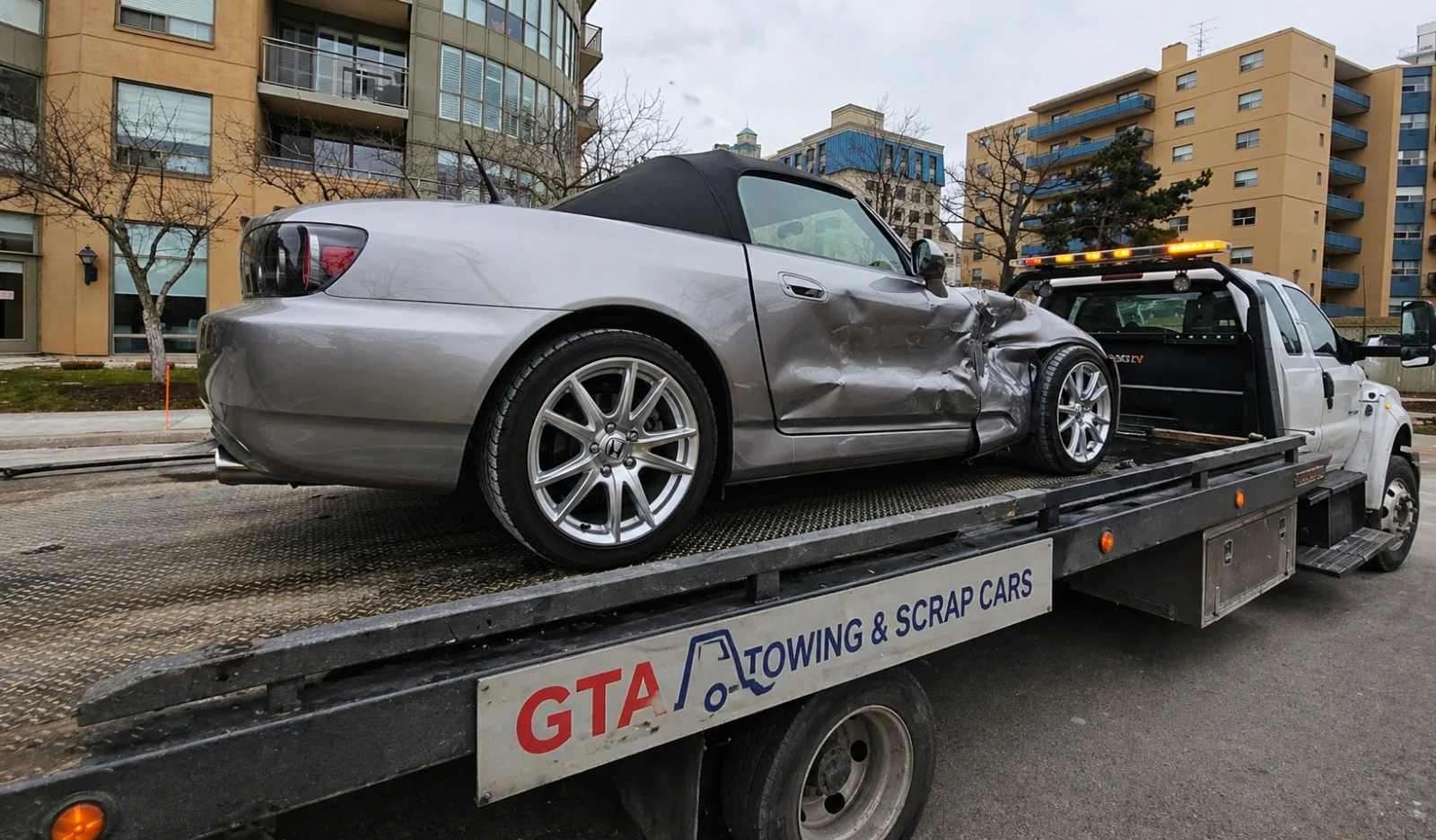 A GTA Towing & Scrap Cars flatbed tow truck secured with recovery straps, actively transporting a weathered, older vehicle for scrap metal recycling.