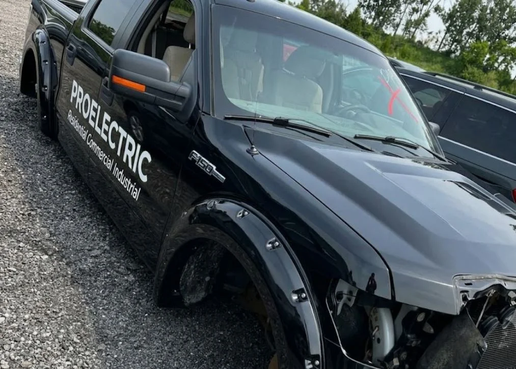 A close-up photograph of a black Ford F-150 truck in a gravel vehicle yard. The truck body and wide fender flares are deep black. On the driver's side, white 'PROELECTRIC Residential Commercial Industrial' text and the 'F-150' badge are visible. The windshield has red 'X' markings, and the front grille and engine compartment are significantly damaged. The truck sits on grey gravel with another dark SUV to the right.