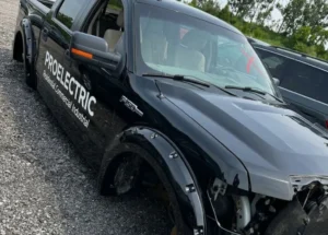 A close-up photograph of a black Ford F-150 truck in a gravel vehicle yard. The truck body and wide fender flares are deep black. On the driver's side, white 'PROELECTRIC Residential Commercial Industrial' text and the 'F-150' badge are visible. The windshield has red 'X' markings, and the front grille and engine compartment are significantly damaged. The truck sits on grey gravel with another dark SUV to the right.